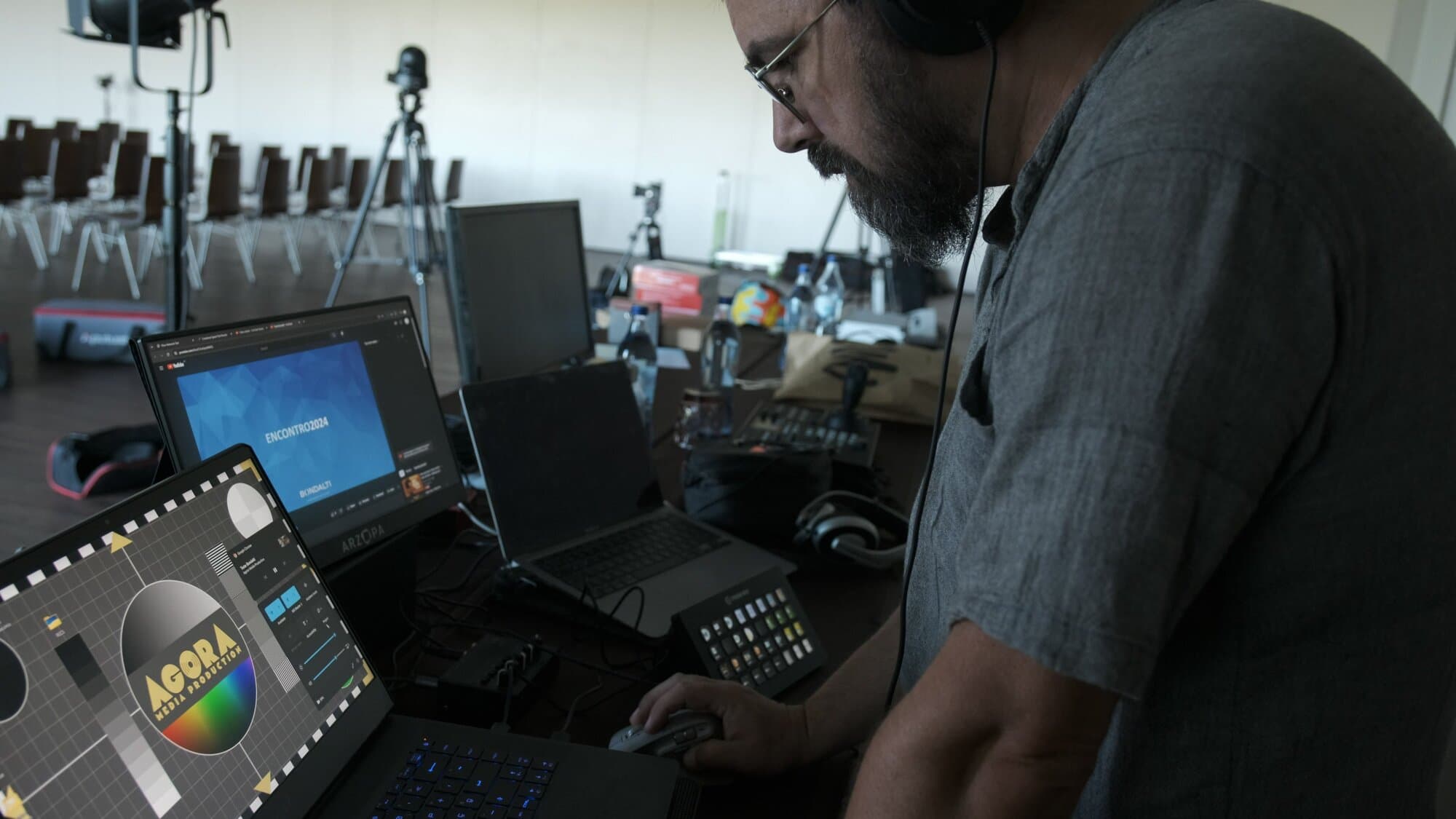 Um técnico da agora na régie, de auscultadores, com a carta de teste da agora no portátil e o cartão do Encontro 2024 no monitor, e a sala ainda vazia ao fundo