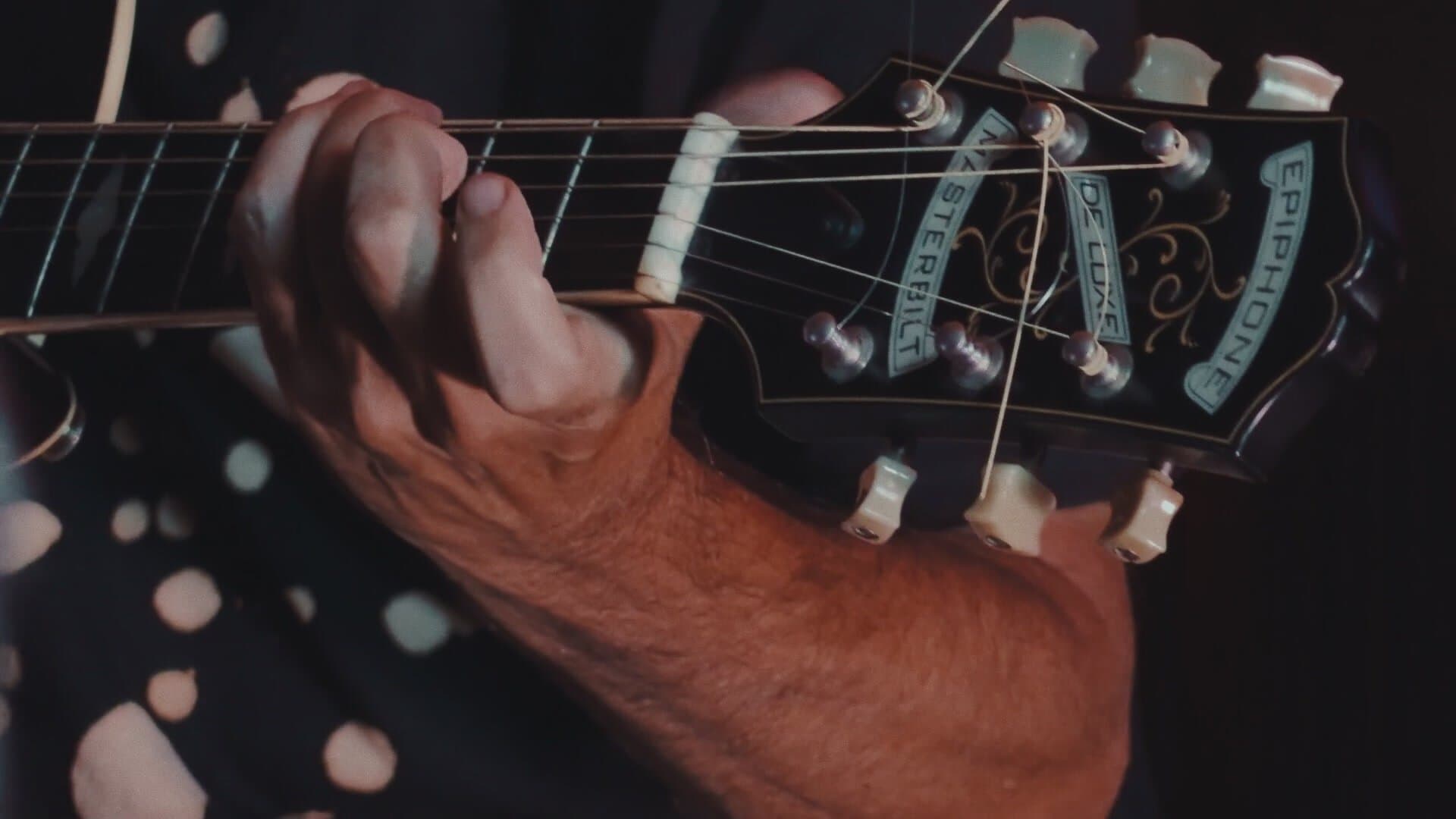 Close-up of Nuno Rebelo's left hand on the guitar neck, next to the headstock of the Epiphone Masterbilt, with his black polka-dot T-shirt behind