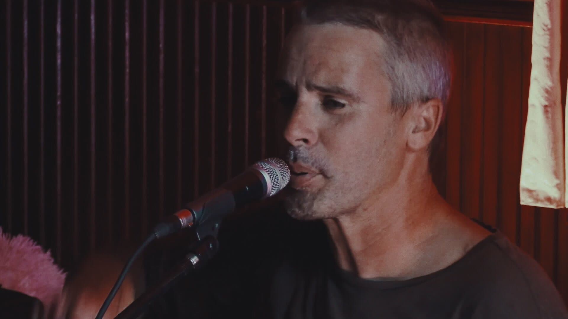 João Leitão singing into the microphone, in close-up, with the curtains behind him