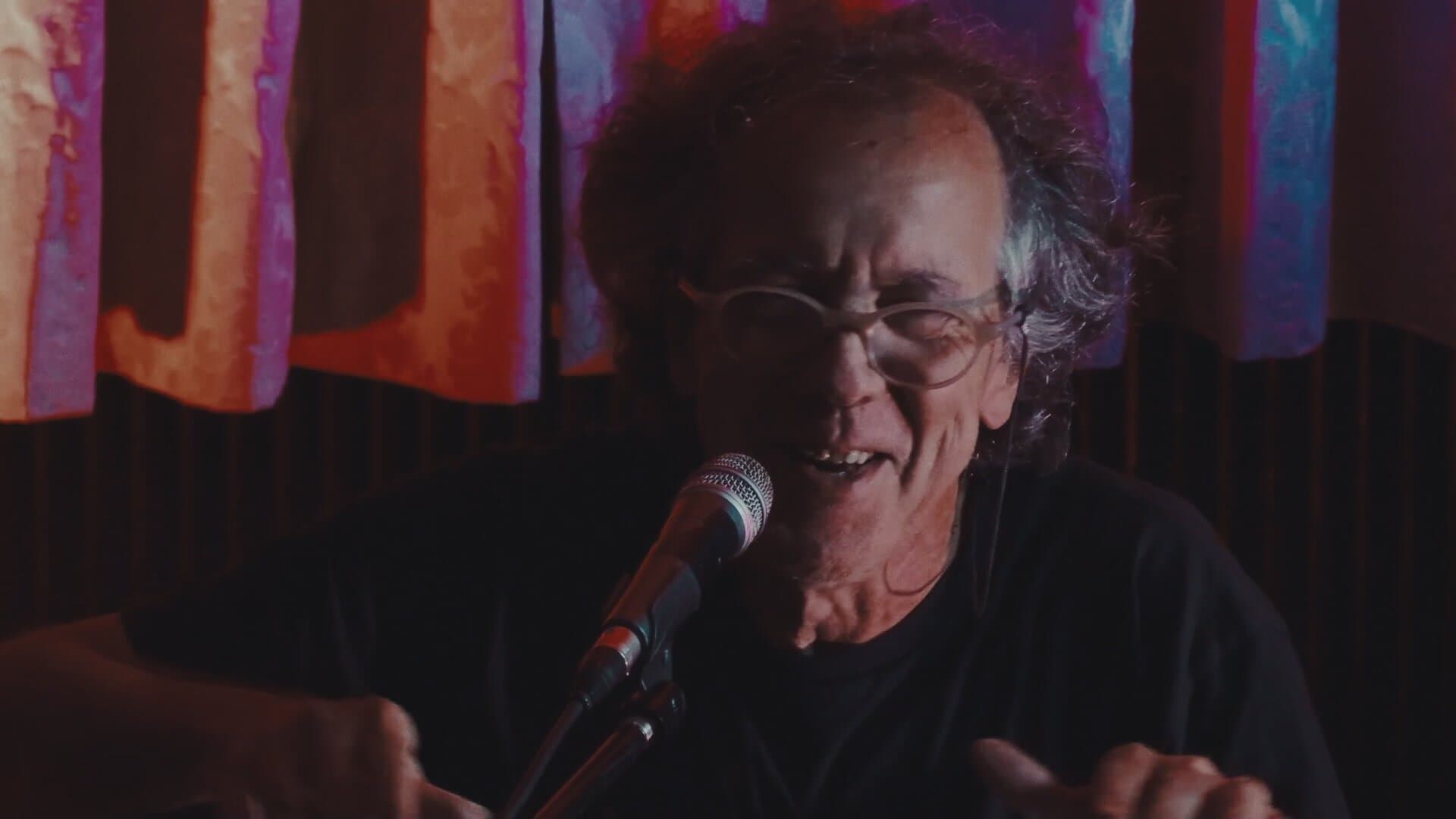 Nuno Rebelo facing the camera at the microphone, smiling at the room, with the curtains behind him, in the same frame before and after colour grading