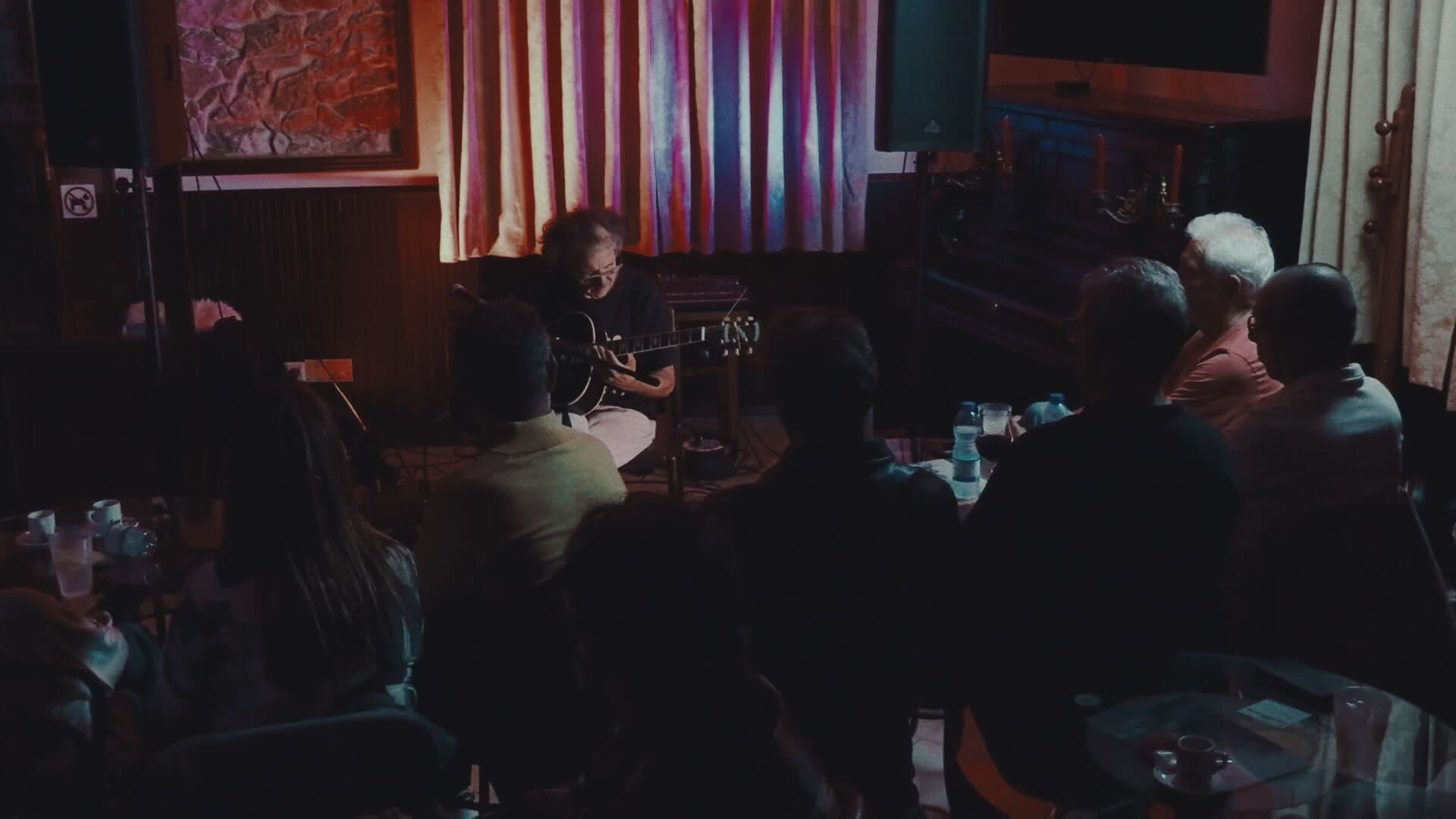 The hall of the Clube Recreativo Penichense during the concert, with the audience at tables, seen from behind, and Nuno Rebelo at the far end, seated with his guitar, in front of the lit curtains