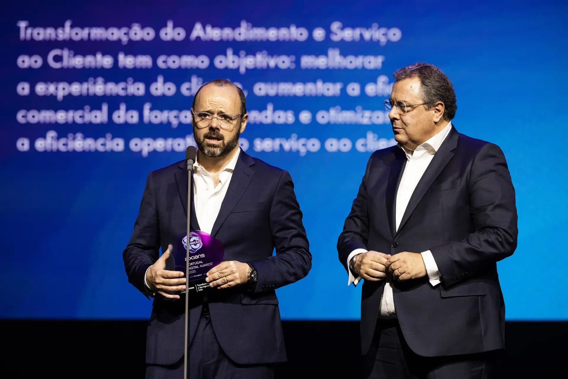 Two Caixa Geral de Depósitos executives at the microphone, one holding the trophy, in front of the screen describing the project