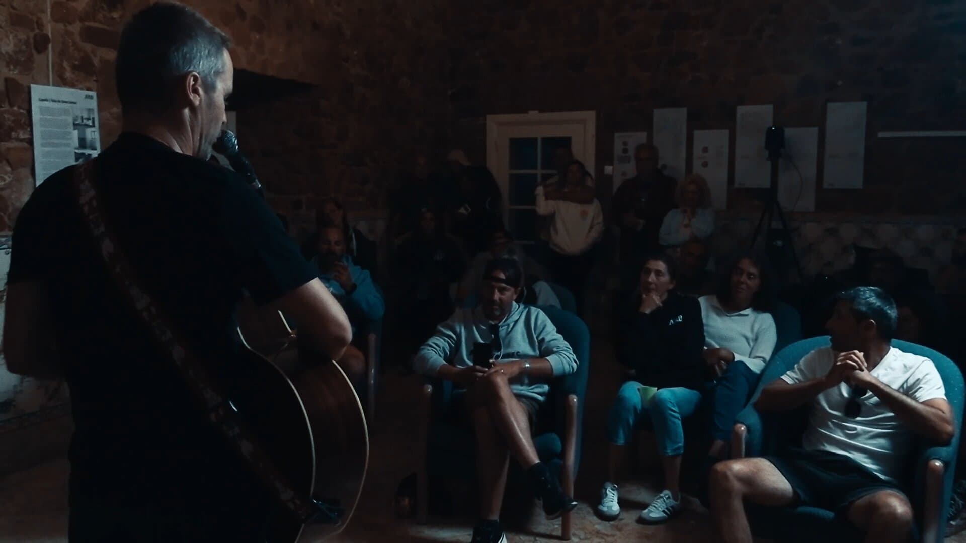 O João Leitão de costas, a cantar para o público sentado em cadeirões, com a janela de portadas fechadas ao fundo