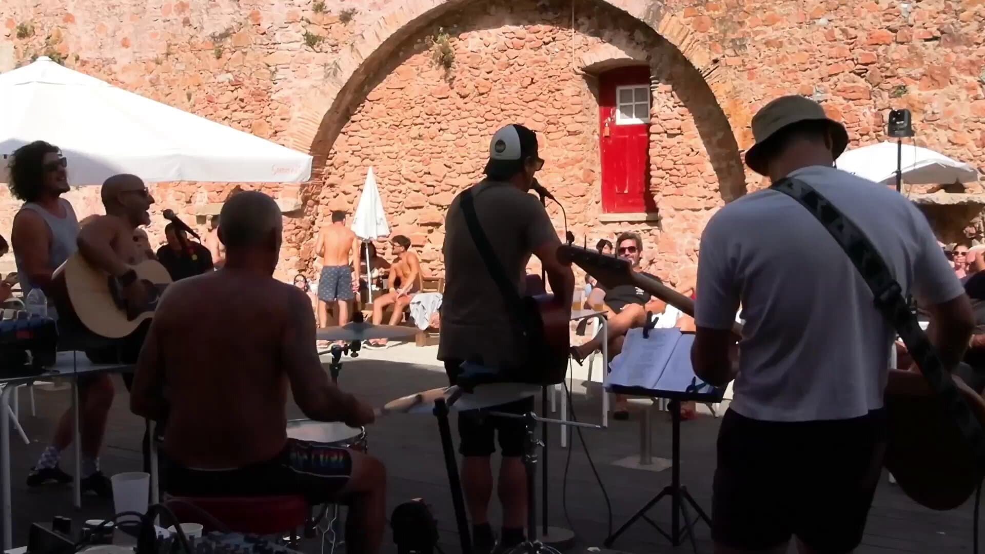 The musicians from behind, playing in the fort's courtyard, with the stone arch, the red door and the audience in the sun in the background