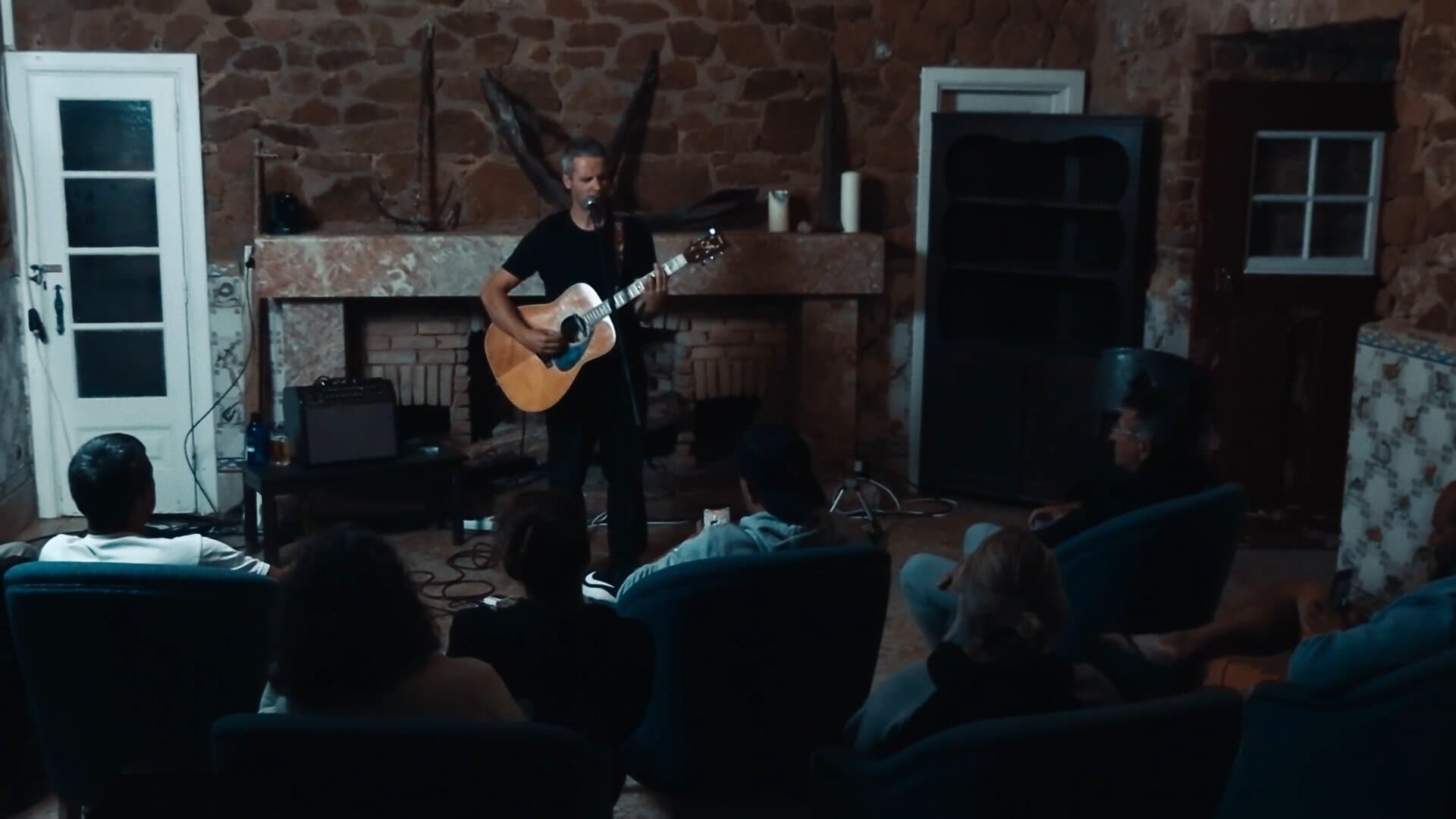 O João Leitão a tocar guitarra diante da lareira do Salão das Princesas, com as paredes de pedra, as portas brancas e o público sentado em primeiro plano